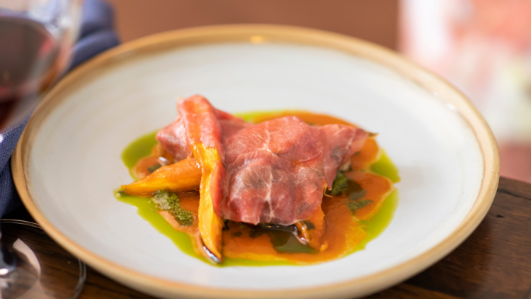 A white plate with lamb belly draped in lamb carpaccio, carrots and parsley oil