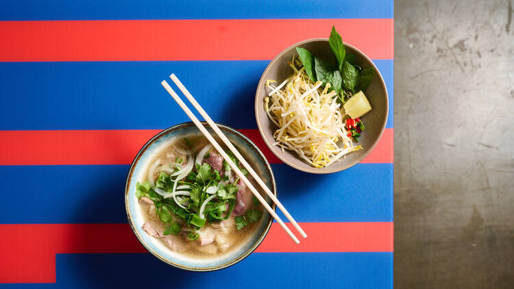 A bowl of beef pho served with a side of bean sprouts and basil.