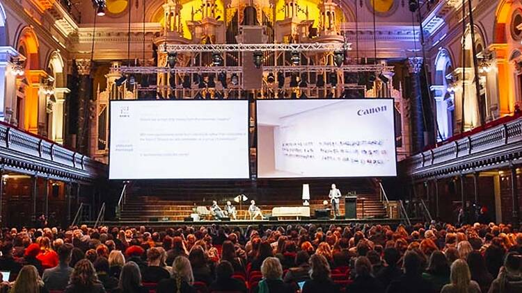 A packed audience in a theatre looks upon a stage with presenters and two big projectors