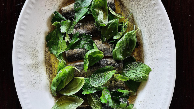 On a white oval plate is pickled sardines garnished with basil and mint leaves