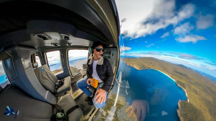 A man sits in a helicopter with no doors, looking down at a beautiful landscape