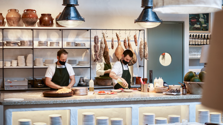 Two chefs stand preparing food on a white counter top, charcuterie hangs behind them