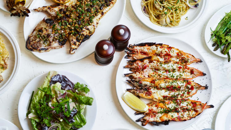 A flat lay of prawns, fish, pasta with clams, and salad on a white table