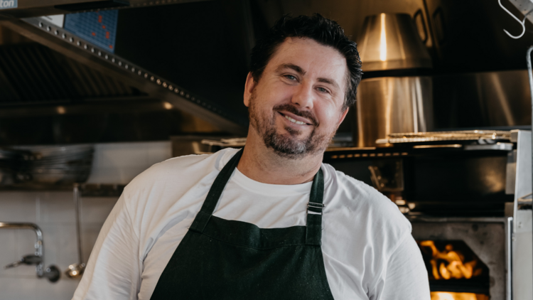 Chef Mike Eggart stands in front of a woodfire oven smiling