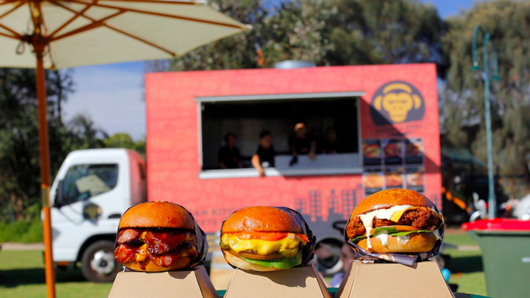 Three burgers in the foreground and a pink food truck in the background.
