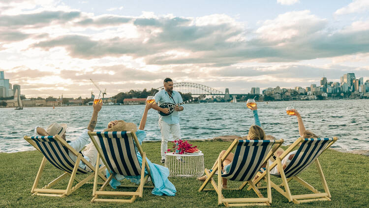 Four people in blue and white deckchairs cheers a man playing a Greek mandolin in front of Sydney Harbour 