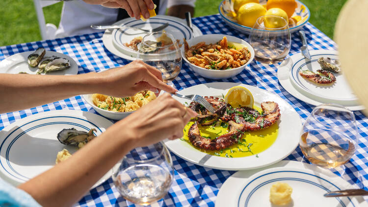 A table covered in a blue and white checked table cloth is covered with Greek food