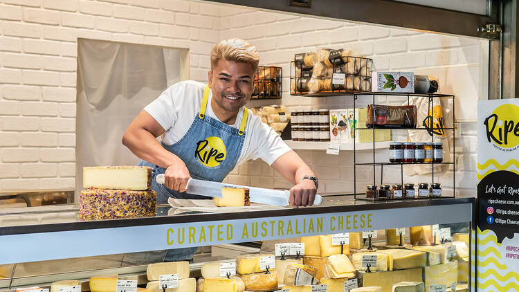 A man stands over a cheese counter and slices through a hunk of cheese with a big knife