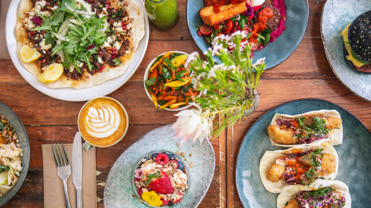 A flat lay of fish tacos, smoothie bowls, manoush, felafel plates with latte on wooden table