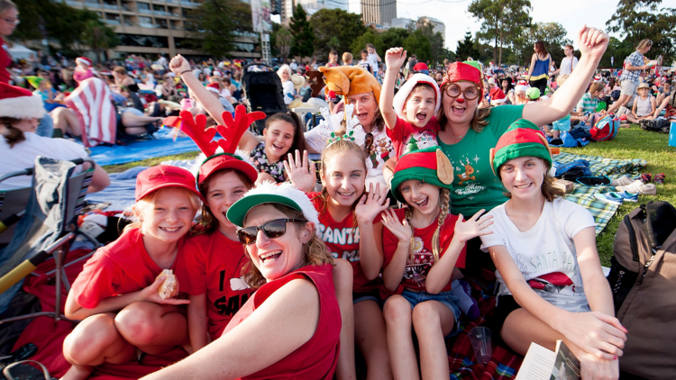 A family is gathered amongst the crowd at a carols event, they are smiling and wearing festive accessories