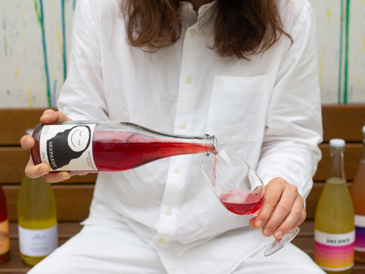 A woman wearing a white collared shirt pours a dark pink pet-nat into a glass