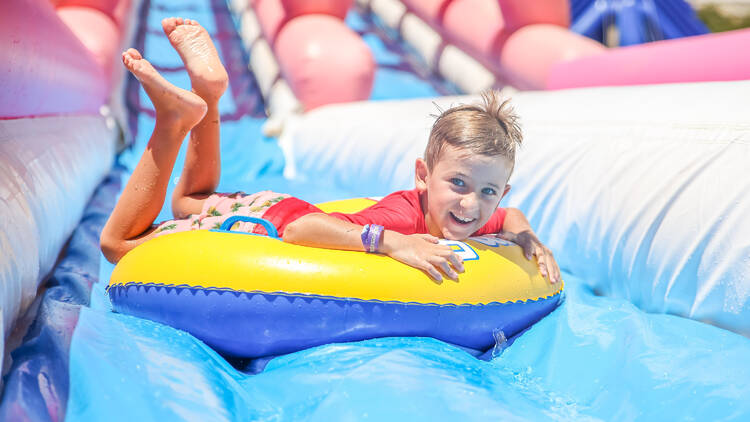 A kid in a bright red rashie smiles at the camera as he rides down a slide on an inflatable ring