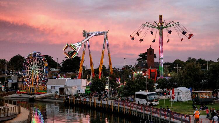 A sunset carnival scene depicting a ferris wheel, giant claw ride and big swing all in action by a riverside location