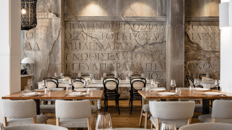 Wooden tables with white and black chairs on a backdrop of sandstone wall with Greek letters