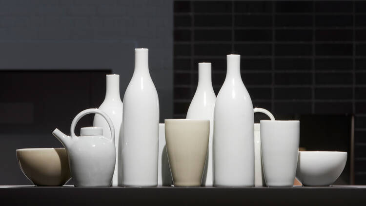 Around 11 white and cream shiny porcelein vessels sitting in a group on a black bench in front of a black tiled background