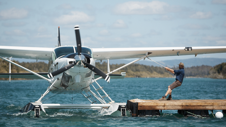 A seaplane is being pulled by hand towards a pontoon by an oyster farmer