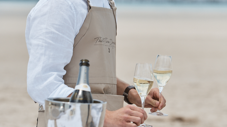 A waiter in a white shirt and brown leather apron holds two glasses of sparkling wine on a sandbank