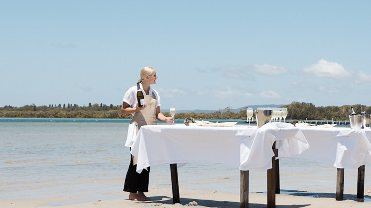 On a sandbank, tables with white linen are dressed with oysters and a waiter pours sparkling wine