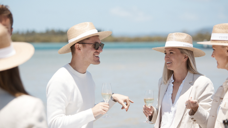 White people are smiling, wearing white and tan suits, drinking sparkling wine