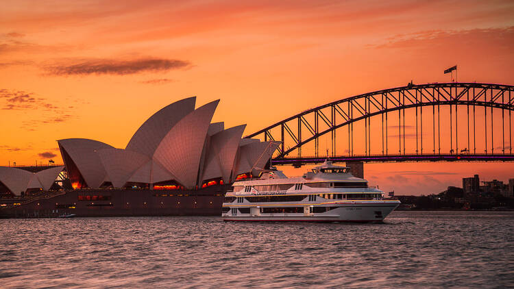 A photograph of a boat in front of the sydney harbour bridge and sydney opera house at sunset