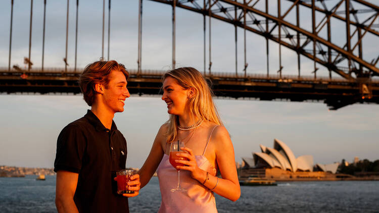 A man and woman standing on the outer deck of a boat at sunset with the sydney harbour bridge towering over them