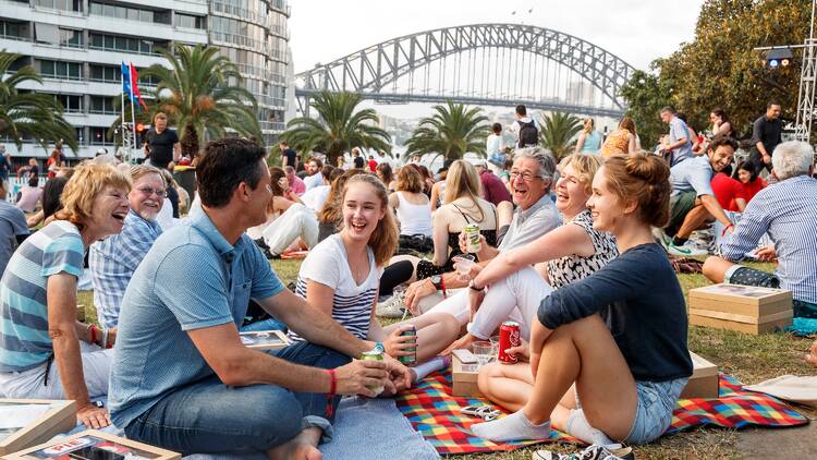 A family having a picnic on the lawn smiling at each other with the harbour bridge in the background