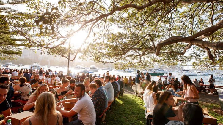 A photograph of a crow of people sitting at picnic tables surrounded by trees 