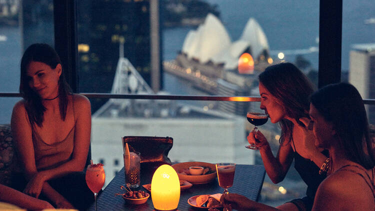 A photo of three women sitting at a window table with drinks and a view of the Sydney opera house