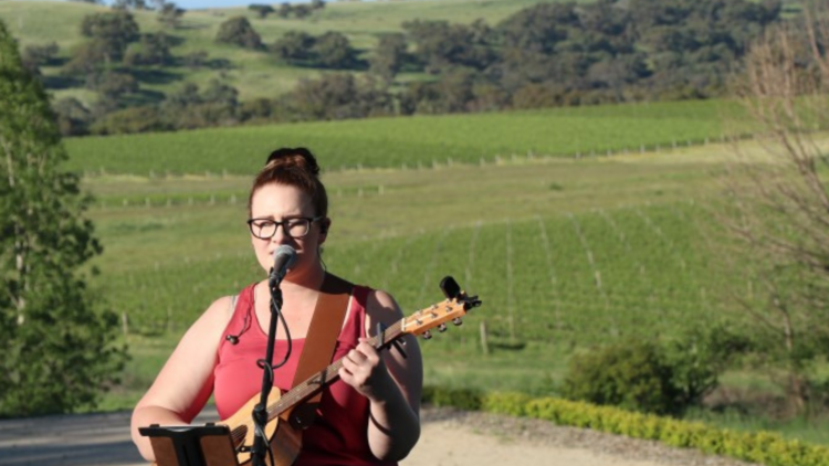 A musician plays her guitar and sings with a vineyard in the background