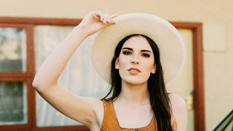 Katie Bates wearing a cowboy hat and leaning against a car. 