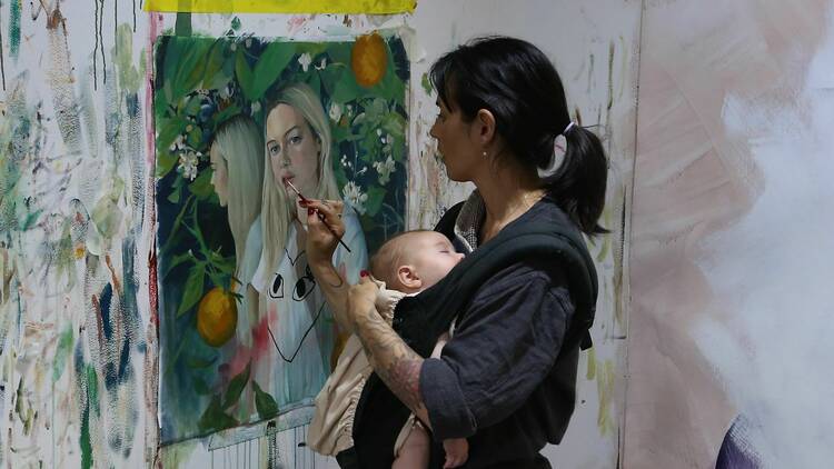 Loretta holding her baby while working on a painting.