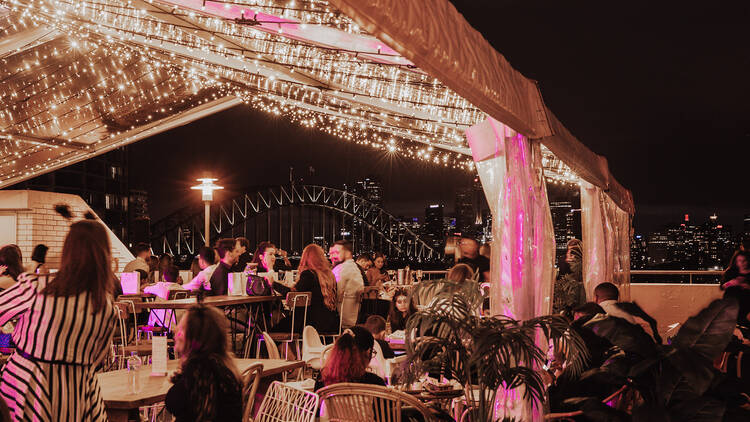 A group of people sit under a fairylight-adorned pagola on a balcony overlooking the harbour