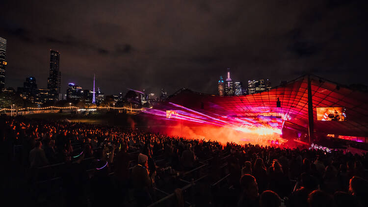 A crowded open-air amphitheatre at night, with pink and red lights plus smoke
