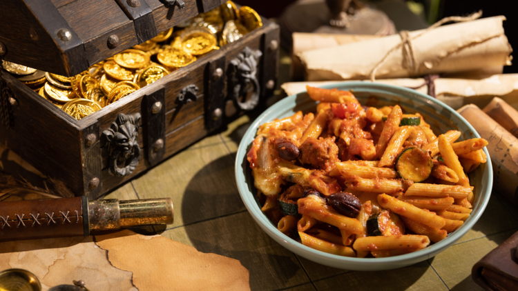 A bowl of penne pasta in tomato sauce on a table with pirate chests and treasure maps