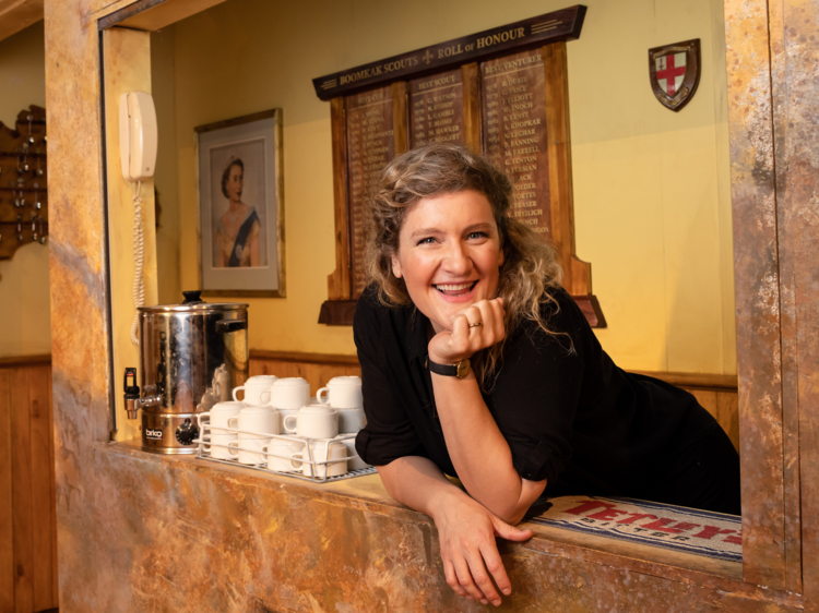 Virginia Gay is posing on the set of the Boomkak Panto. She wears all black and leans out the window of a canteen next to a coffee dispender, mugs, a portrait of the queen and a spoon collection on an Australia-shaped hanger.