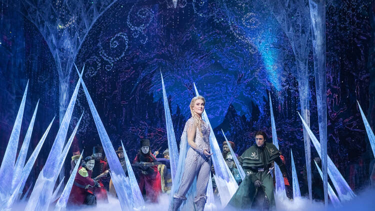 A blond woman in a pale blue jumpsuit stands on a stage surrounded by icicles