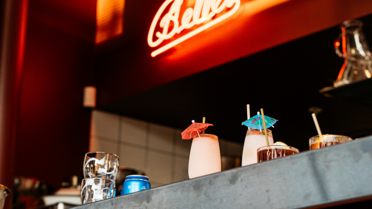 A bar with cocktails lined up and a neon sign reading Belles
