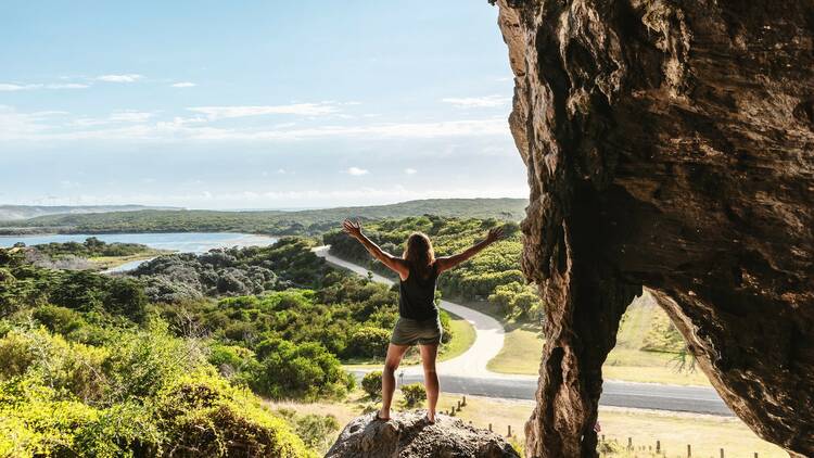 A person stands on a boulder with their arms outstretched at the entrance to Tarragal Caves. In the distance you can see forest and a large body of water