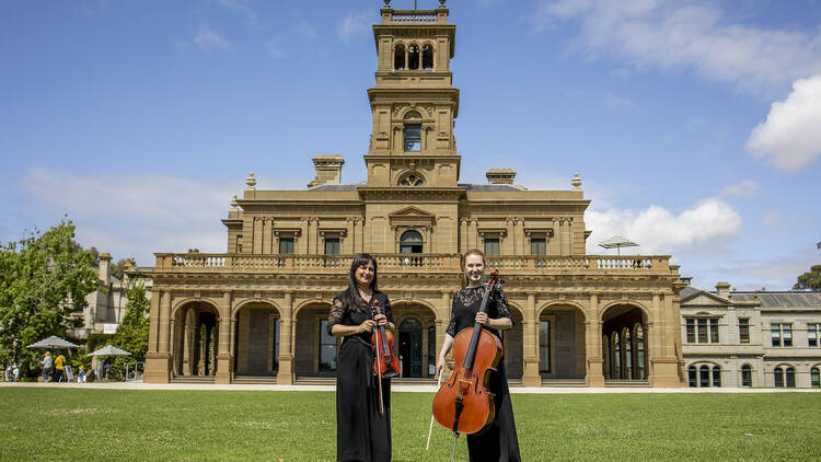 Two women standing in front of Werribee Mansion while holding string instruments.