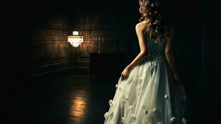 A woman in a white ball gown stands in front of a fancy chandelier