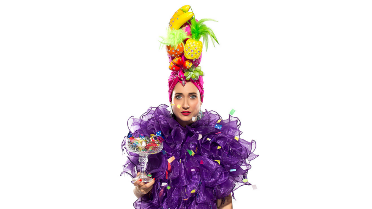 Thalia-Joan wears a frilly purple garment, a fruit headpiece and holds a large cocktail glass filled with confetti