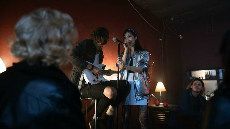 A woman singing into a micrphone while a man is seated next to her playing the guitar.