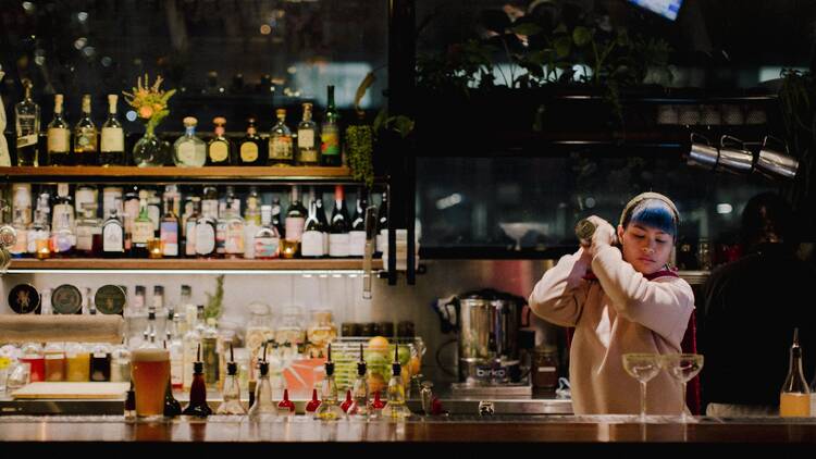 A woman shaking a cocktail behind a bar, and in the background is a wall filled with spirits. 