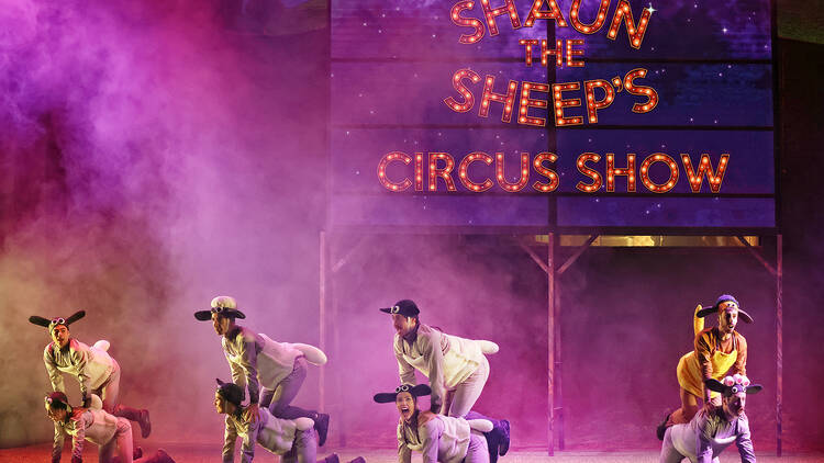 Eight performers dressed as sheep pose on stage in front of a neon sign that says 'Shaun the Sheep's Circus Show'