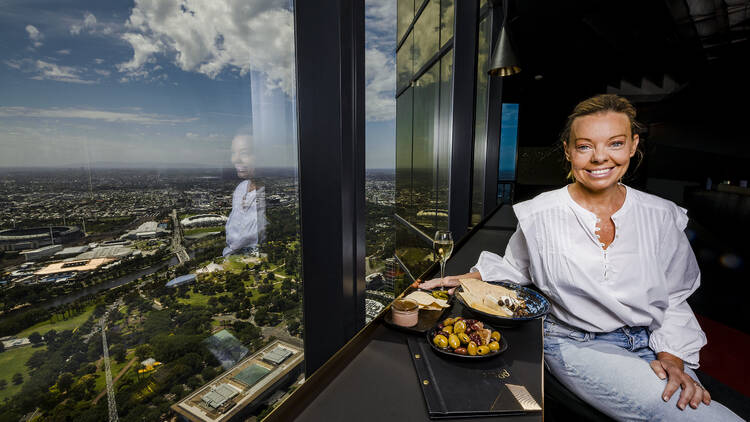 A woman sitting inside of the Melbourne Skydeck with a plate of food and glass of wine.