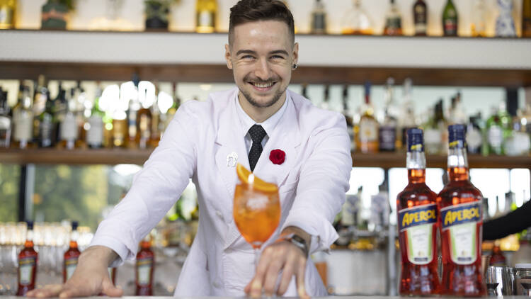 A smiling bartender pushes and Aperol Spritz across the bar towards the camera