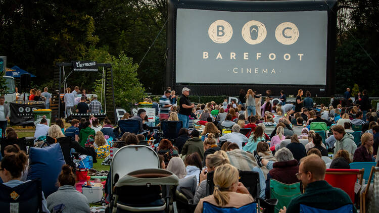 A crowd of people facing away from the camera and towards a large outdoor screen that reads 'Barefoot Cinema'. 