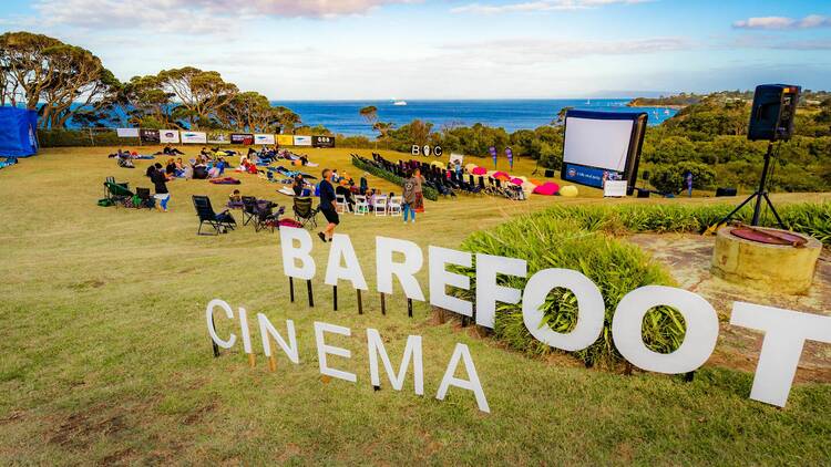 A lawn with a sign that reads 'Barefoot Cinema' and a crowd of people visible in the background, sitting before a large screen.
