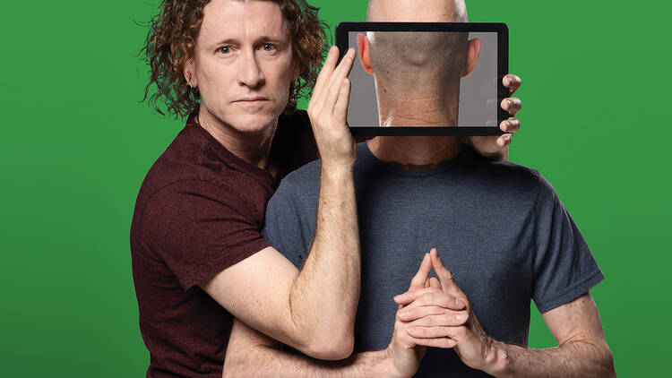 A curly haired man holds an iPad screen up against a the face of a bald man