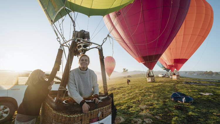 A woman stands in the basket of a hot air balloon, while more hot air balloons prepare for take-off in the background